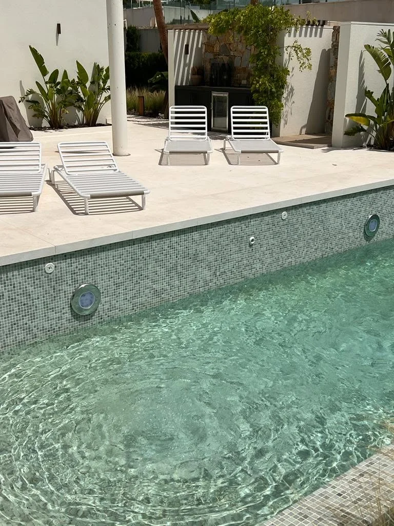 Green mosaic swimming pool beside a pale terrace and white sun loungers